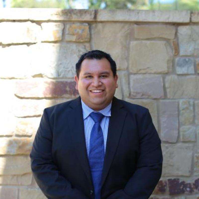 Headshot of a man smiling wearing a navy blue suit and tie standing in front of a stone wall.