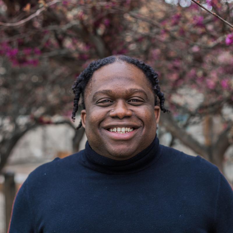 Headshot of a smiling man wearing a blue long sleeve shirt, against a tree background.