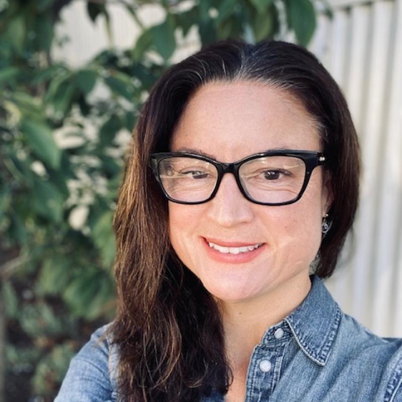 Selfie of a smiling person with shoulder-length brown hair, wearing a denim top.